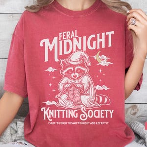 Puede incluir: Camiseta de color carmes&iacute; con el texto "FERAL MIDNIGHT KNITTING SOCIETY". El gr&aacute;fico presenta un mapache con gorro de dormir, tejiendo. Tambi&eacute;n se incluye el texto "I SAID I'D FINISH THIS WIP TONIGHT AND I MEANT IT".