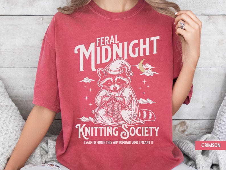 Puede incluir: Camiseta de color carmes&iacute; con el texto "FERAL MIDNIGHT KNITTING SOCIETY". El gr&aacute;fico presenta un mapache con gorro de dormir, tejiendo. Tambi&eacute;n se incluye el texto "I SAID I'D FINISH THIS WIP TONIGHT AND I MEANT IT".
