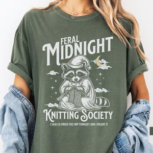 Puede incluir: Camiseta verde musgo con un dise&ntilde;o de un mapache tejiendo, con gorro de dormir, y las palabras "Feral Midnight Knitting Society". El texto "I said I'd finish this WIP tonight and I meant it" est&aacute; debajo.