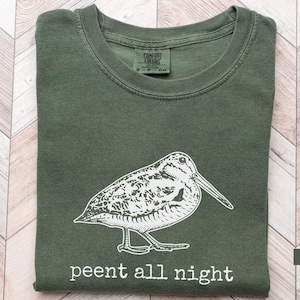 May include: A moss green t-shirt with a white illustration of a bird and the text "peent all night." The shirt is folded and has a crew neck. The Comfort Colors tag is visible at the top. The shirt is made of a soft material.