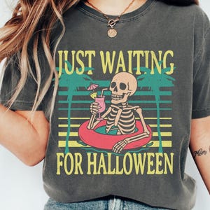 Peut inclure: T-shirt gris fonc&eacute; avec l'inscription &laquo; JUST WAITING FOR HALLOWEEN &raquo;. Le motif repr&eacute;sente un squelette dans une bou&eacute;e avec un cocktail, des palmiers et des lignes horizontales jaunes, vertes et rouges.