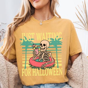 Peut inclure: T-shirt moutarde avec l'inscription "JUST WAITING FOR HALLOWEEN". Le graphisme repr&eacute;sente un squelette dans une bou&eacute;e avec une boisson. Des palmiers et un motif de coucher de soleil sont &eacute;galement inclus.