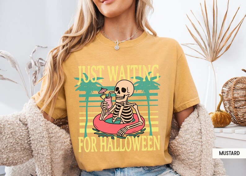 Peut inclure: T-shirt moutarde avec l'inscription "JUST WAITING FOR HALLOWEEN". Le graphisme repr&eacute;sente un squelette dans une bou&eacute;e avec une boisson. Des palmiers et un motif de coucher de soleil sont &eacute;galement inclus.
