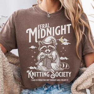 Puede incluir: Camiseta marr&oacute;n con un dise&ntilde;o de un mapache con gorro de dormir tejiendo. El texto dice "Feral Midnight Knitting Society" y "I said I'd finish this WIP tonight and I meant it."