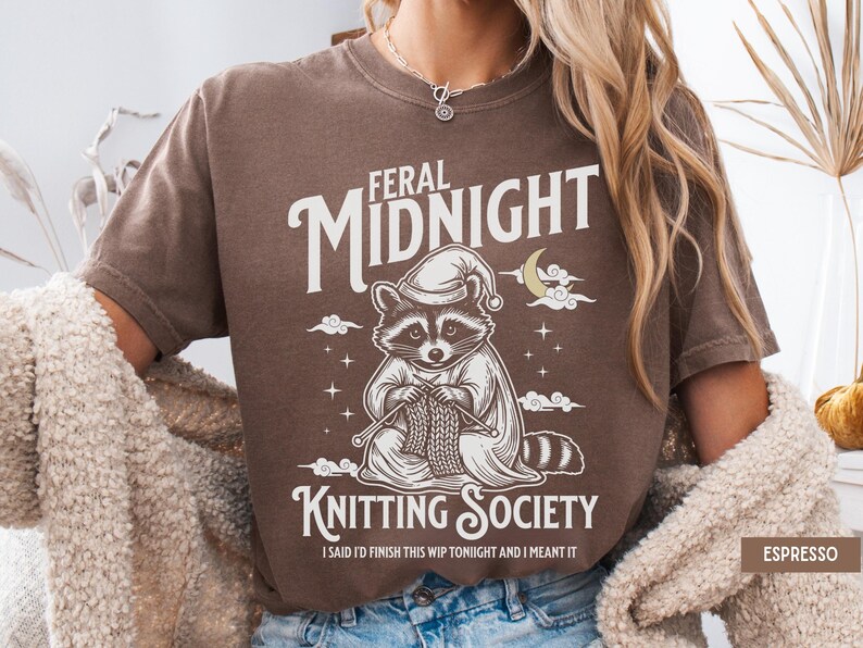 Puede incluir: Camiseta marr&oacute;n con un dise&ntilde;o de un mapache con gorro de dormir tejiendo. El texto dice "Feral Midnight Knitting Society" y "I said I'd finish this WIP tonight and I meant it."