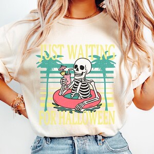Peut inclure: T-shirt ivoire avec un motif de squelette dans une bou&eacute;e rose, tenant un cocktail. Le texte "JUST WAITING FOR HALLOWEEN" est en jaune. Le design comprend des palmiers et des lignes horizontales.