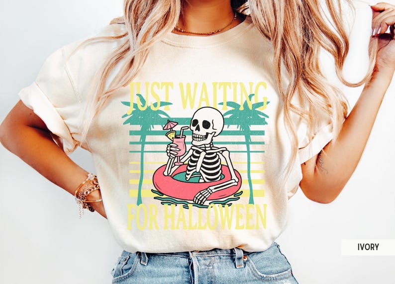 Peut inclure: T-shirt ivoire avec un motif de squelette dans une bou&eacute;e rose, tenant un cocktail. Le texte "JUST WAITING FOR HALLOWEEN" est en jaune. Le design comprend des palmiers et des lignes horizontales.
