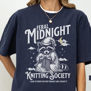 Puede incluir: Camiseta azul marino con un mapache con gorro de dormir, tejiendo. La camiseta presenta el texto "Feral Midnight Knitting Society" y "I said I'd finish this WIP tonight and I meant it."
