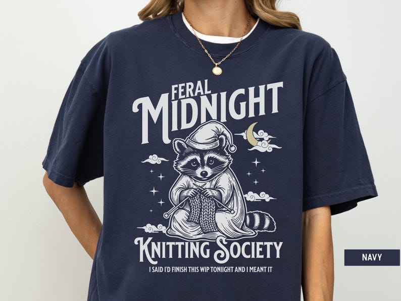 Puede incluir: Camiseta azul marino con un mapache con gorro de dormir, tejiendo. La camiseta presenta el texto "Feral Midnight Knitting Society" y "I said I'd finish this WIP tonight and I meant it."