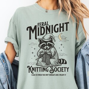 Puede incluir: Una camiseta verde claro con las palabras "FERAL MIDNIGHT KNITTING SOCIETY" en negro. La camiseta presenta un mapache con gorro de dormir tejiendo. El texto "I SAID I'D FINISH THIS WIP TONIGHT AND I MEANT IT" est&aacute; debajo.