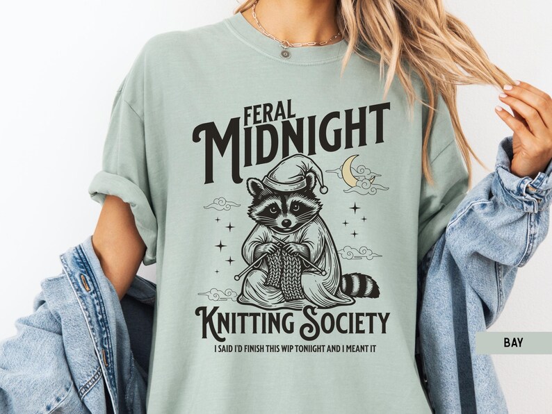 Puede incluir: Una camiseta verde claro con las palabras "FERAL MIDNIGHT KNITTING SOCIETY" en negro. La camiseta presenta un mapache con gorro de dormir tejiendo. El texto "I SAID I'D FINISH THIS WIP TONIGHT AND I MEANT IT" est&aacute; debajo.