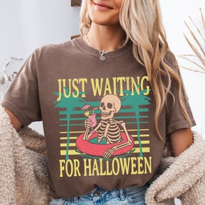 Peut inclure: T-shirt marron avec l'inscription &laquo; JUST WAITING FOR HALLOWEEN &raquo;. Le motif repr&eacute;sente un squelette dans une bou&eacute;e avec un cocktail, sur fond de palmiers et de coucher de soleil.