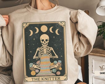 May include: Sand-colored sweatshirt featuring a tarot card design. The card depicts a skeleton knitting with colorful yarn, surrounded by moons and stars. The text "THE KNITTER" is at the bottom. A cozy and unique apparel item.