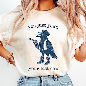 Puede incluir: Camiseta color marfil con un cuervo de dibujos animados azul marino con sombrero de vaquero, chaleco y botas, sosteniendo un revólver. El texto "you just yee'd" y "your last caw" está impreso arriba y abajo.