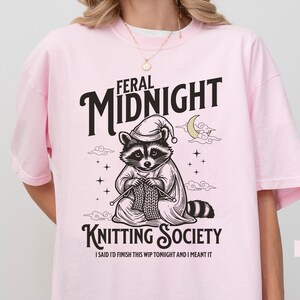 Puede incluir: Camiseta rosa claro con un dise&ntilde;o de un mapache con gorro de dormir tejiendo. El texto dice "FERAL MIDNIGHT KNITTING SOCIETY" con la frase "I SAID I'D FINISH THIS WIP TONIGHT AND I MEANT IT" debajo. La camiseta est&aacute; etiquetada como "BLOSSOM".