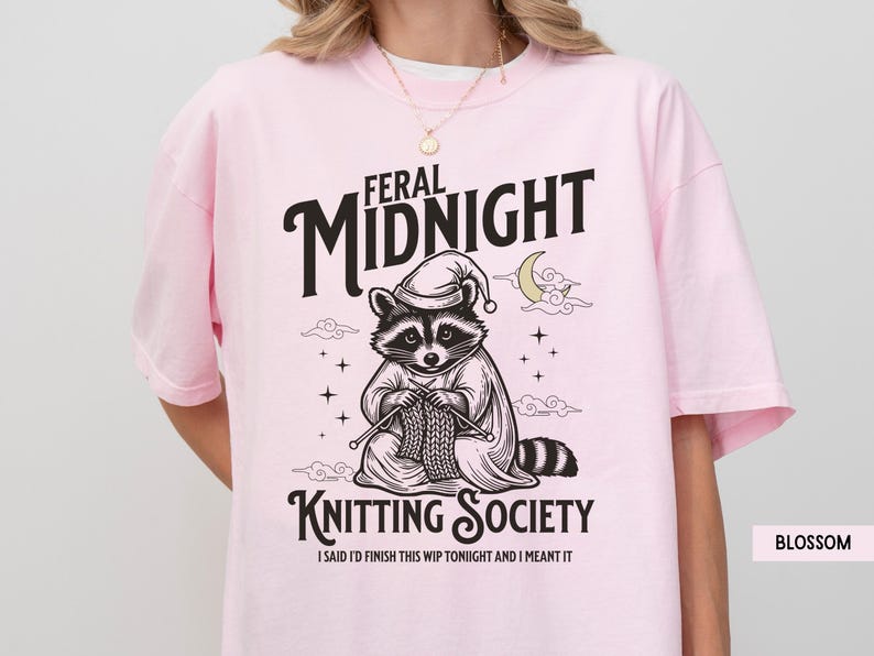 Puede incluir: Camiseta rosa claro con un dise&ntilde;o de un mapache con gorro de dormir tejiendo. El texto dice "FERAL MIDNIGHT KNITTING SOCIETY" con la frase "I SAID I'D FINISH THIS WIP TONIGHT AND I MEANT IT" debajo. La camiseta est&aacute; etiquetada como "BLOSSOM".
