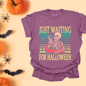 Peut inclure: T-shirt de couleur baie avec un motif de squelette dans une bou&eacute;e, tenant une boisson. Le texte dit "JUST WAITING FOR HALLOWEEN". Des citrouilles, des araign&eacute;es et des chauves-souris sont dispos&eacute;es autour du t-shirt, cr&eacute;ant un th&egrave;me d'Halloween.