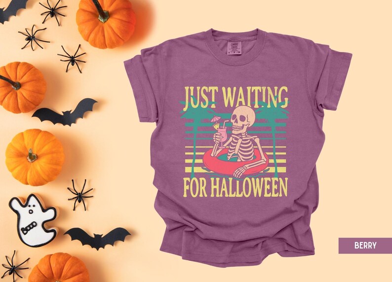 Peut inclure: T-shirt de couleur baie avec un motif de squelette dans une bou&eacute;e, tenant une boisson. Le texte dit "JUST WAITING FOR HALLOWEEN". Des citrouilles, des araign&eacute;es et des chauves-souris sont dispos&eacute;es autour du t-shirt, cr&eacute;ant un th&egrave;me d'Halloween.