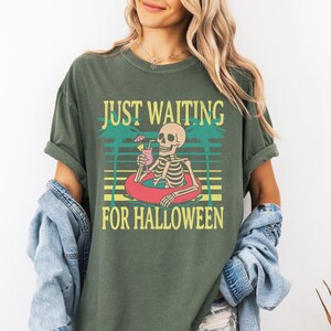 Peut inclure: T-shirt vert mousse avec un motif d'un squelette dans une bou&eacute;e, tenant un cocktail. Le texte "JUST WAITING FOR HALLOWEEN" est au-dessus et en dessous du squelette. Le t-shirt a des manches courtes et une coupe d&eacute;contract&eacute;e.