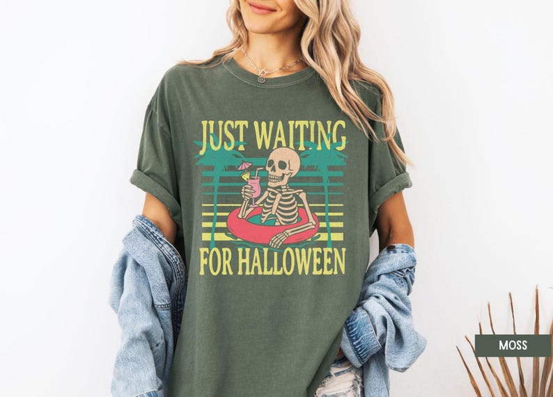 Peut inclure: T-shirt vert mousse avec un motif d'un squelette dans une bou&eacute;e, tenant un cocktail. Le texte "JUST WAITING FOR HALLOWEEN" est au-dessus et en dessous du squelette. Le t-shirt a des manches courtes et une coupe d&eacute;contract&eacute;e.