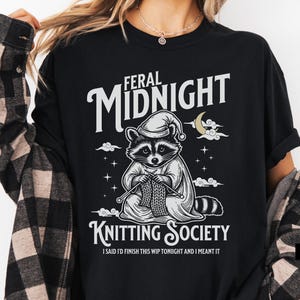 Puede incluir: Camiseta negra con un mapache con gorro de dormir, tejiendo. El texto dice "FERAL MIDNIGHT KNITTING SOCIETY" con la frase "I SAID I'D FINISH THIS WIP TONIGHT AND I MEANT IT".