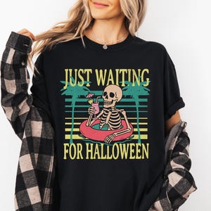 Peut inclure: T-shirt noir avec un motif d'un squelette dans une bou&eacute;e, tenant un cocktail. Le texte dit "JUST WAITING FOR HALLOWEEN" en jaune et vert. Un design amusant et saisonnier.