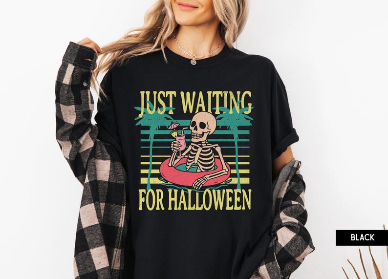 Peut inclure: T-shirt noir avec un motif d'un squelette dans une bou&eacute;e, tenant un cocktail. Le texte dit "JUST WAITING FOR HALLOWEEN" en jaune et vert. Un design amusant et saisonnier.