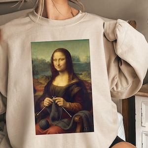 Puede incluir: Una sudadera de cuello redondo color arena con un estampado gráfico de la Mona Lisa tejiendo con lana y agujas grises. La sudadera tiene mangas largas, cuello acanalado y un corte relajado. La palabra SAND está impresa en la esquina inferior.