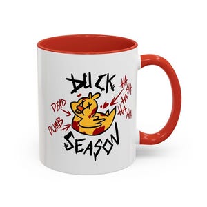 Puede incluir: Taza de cerámica blanca con interior y asa rojos. La taza presenta un pato de dibujos animados con salpicaduras de sangre y las palabras "Duck Season" en negro, con fuente manuscrita. El texto adicional incluye "Dead" y "Dumb".