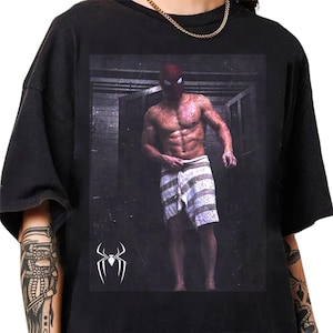 May include: A black short-sleeved t-shirt featuring a graphic print of a muscular figure wearing a red mask and a white and gray striped towel. The figure stands in a dark, tiled room. A white spider logo is positioned in the lower left corner.