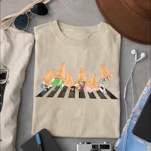 May include: Beige t-shirt featuring a cartoon design of characters crossing a crosswalk under orange traffic cones. The characters include a frog, a pig, and Buzz Lightyear. The shirt is made of a soft material.