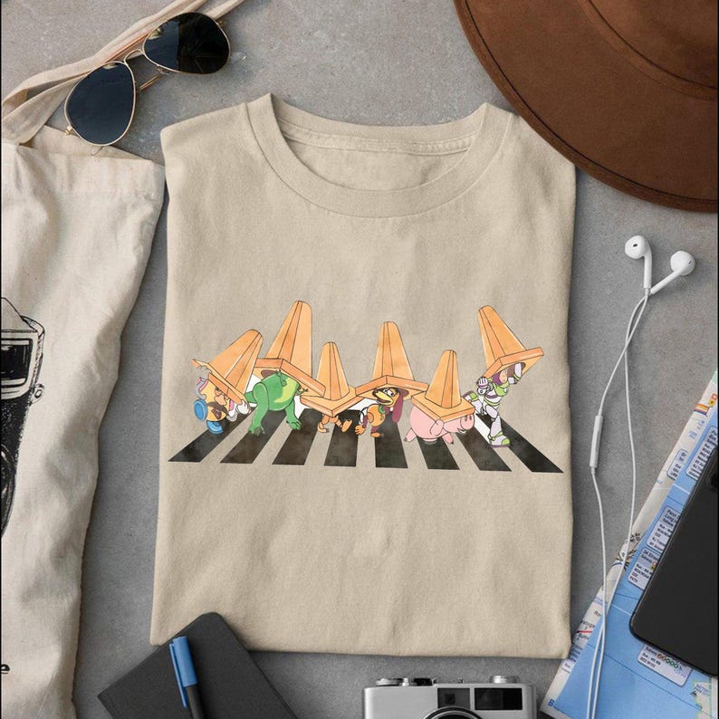 May include: Beige t-shirt featuring a cartoon design of characters crossing a crosswalk under orange traffic cones. The characters include a frog, a pig, and Buzz Lightyear. The shirt is made of a soft material.