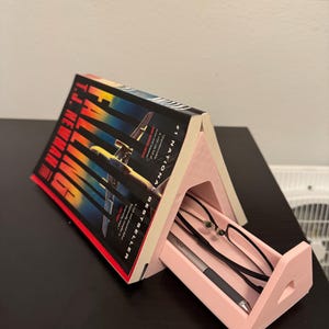 May include: A pink 3D printed book stand with a drawer that can hold glasses and a pen. The stand is holding a paperback book titled "Fatal  Flight" by  J.T.  McKinley. The book is a #1 National Bestseller.