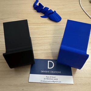 May include: Two 3D printed objects, one black and one blue, on a wooden surface. A business card with the text "Drivebye Creations" is in the center of the image.