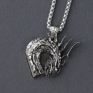 Puede incluir: Un collar de plata con un colgante de dragón. El dragón está enroscado en un círculo, con la cabeza y la cola tocándose. El colgante es detallado e intrincado.