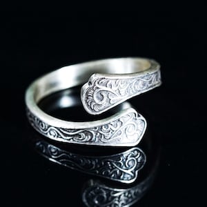 May include: A silver-colored adjustable ring with an ornate, swirling floral pattern. The ring has an open design, with the ends overlapping. The ring is displayed on a reflective black surface.