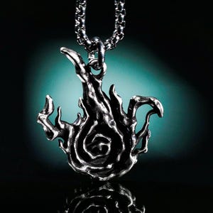 May include: A silver-colored pendant necklace featuring a stylized flame design. The pendant has a detailed, textured surface and hangs from a matching chain. The flame design has a swirling center and pointed edges.