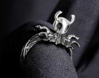 May include: A silver ring featuring a detailed sculpture of a knight with a helmet and wings. The ring is designed to be worn on the finger and has a textured band.