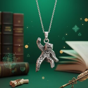 Pu&ograve; includere: Una collana con pendente in argento raffigurante una figura stilizzata con intricati motivi geometrici. Il pendente &egrave; appeso a una delicata catena. Lo sfondo include libri, pergamene e una penna, suggerendo un tema letterario o fantasy.