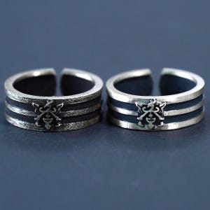 May include: Two silver adjustable rings. Each ring has a dark band between two silver bands, with a decorative emblem on the front. The rings are set against a dark background, highlighting their intricate design and metallic finish.