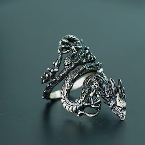 May include: A detailed silver dragon ring. The dragon wraps around the finger, with intricate scales and a fierce expression. The ring is set against a dark background, highlighting the metallic details.