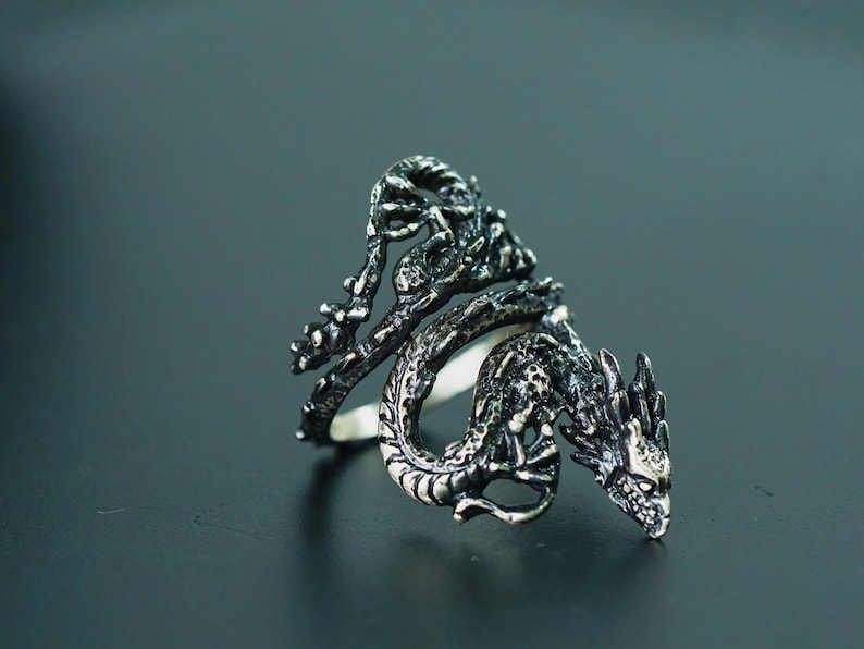 May include: A detailed silver dragon ring. The dragon wraps around the finger, with intricate scales and a fierce expression. The ring is set against a dark background, highlighting the metallic details.