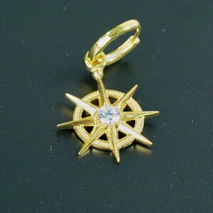 K&ouml;nnte beinhalten: Goldener Ohrring mit einem kompassartigen Design, das einen klaren Kristall in der Mitte tr&auml;gt. Der Ohrring hat einen B&uuml;gelverschluss.