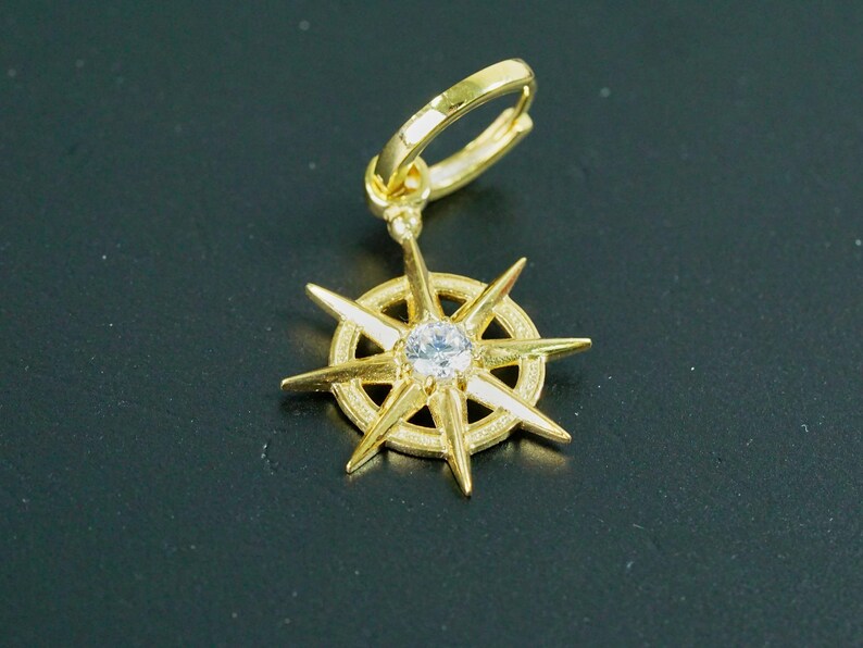 K&ouml;nnte beinhalten: Goldener Ohrring mit einem kompassartigen Design, das einen klaren Kristall in der Mitte tr&auml;gt. Der Ohrring hat einen B&uuml;gelverschluss.
