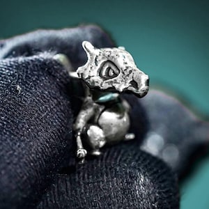 May include: A small, silver-toned metal figurine shaped like a creature with a skull-like head and a rounded body. The surface of the metal has a textured, weathered appearance with visible indentations and a metallic sheen throughout the piece.