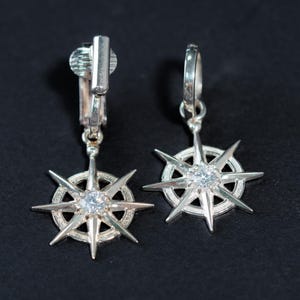 K&ouml;nnte beinhalten: Ein Paar silberne Ohrclips mit einem Sternburst-Design und einem klaren Kristall in der Mitte.