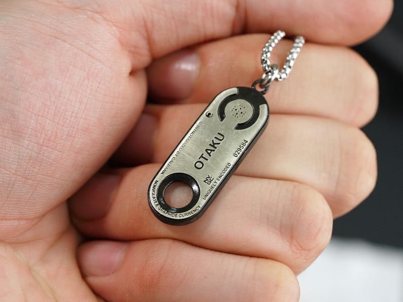May include: A silver and black pendant necklace with the word "OTAKU" engraved on it. The pendant is shaped like a small rectangle with rounded edges and has a small circle in the center. The necklace is on a silver chain.