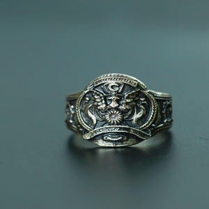 May include: A silver ring with a detailed design featuring a winged creature and a star. The ring has a black background and gold accents.