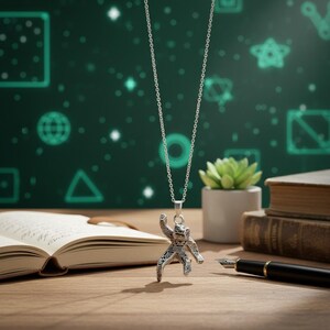 Pu&ograve; includere: Collana con pendente in argento con un design dettagliato di astronauta. Il pendente &egrave; appeso a una catena delicata. Lo sfondo include un libro, una penna e una piccola pianta in vaso, il tutto su una superficie di legno.
