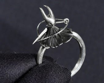 May include: A silver ring featuring a detailed sculpture of a knight with a sword and a cape. The knight is wearing a helmet and holding a shield.
